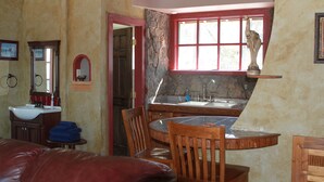 A dining area with a round table, chairs, a red-framed window, a stone wall, and a sink with a mirror.