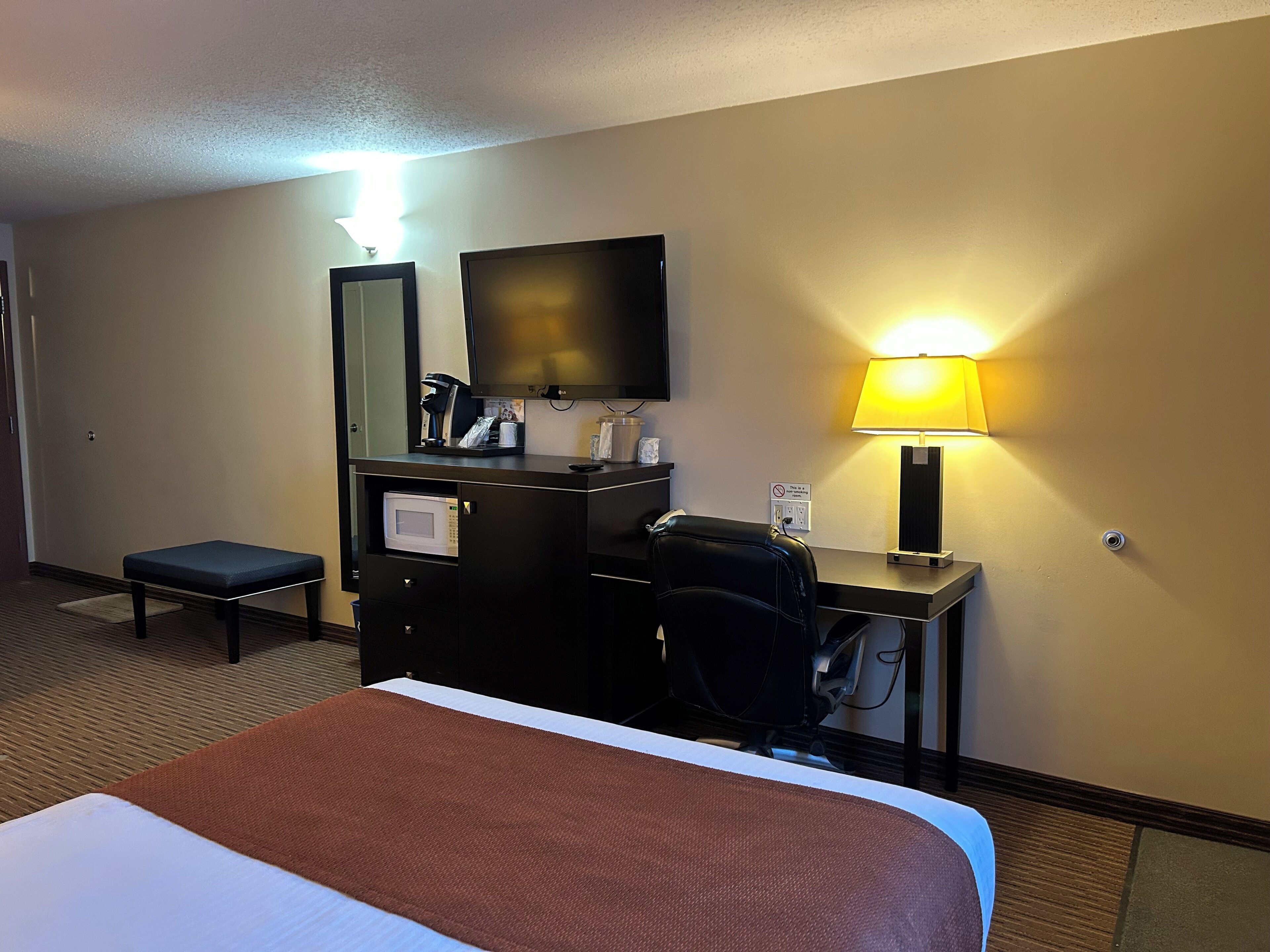 Une chambre d’hôtel équipée d’un lit, d’un bureau avec un ordinateur, d’une chaise, d’une télévision, d’une lampe et d’un four à micro-ondes.