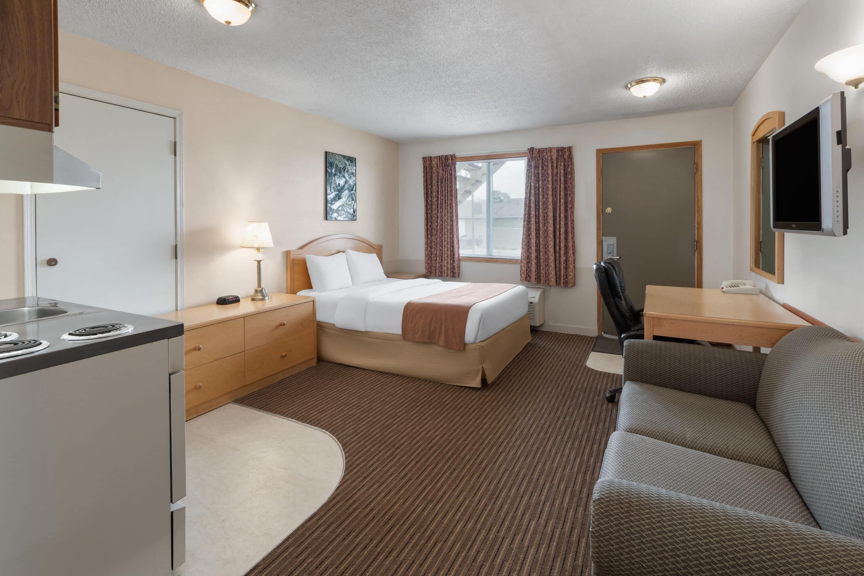 A hotel room with a bed, a desk, a chair, a TV, and a kitchenette.