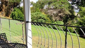 Un espace extérieur abrité, avec des chaises et une table en métal, donnant sur une zone verdoyante agrémentée d’arbres et d’une clôture.
