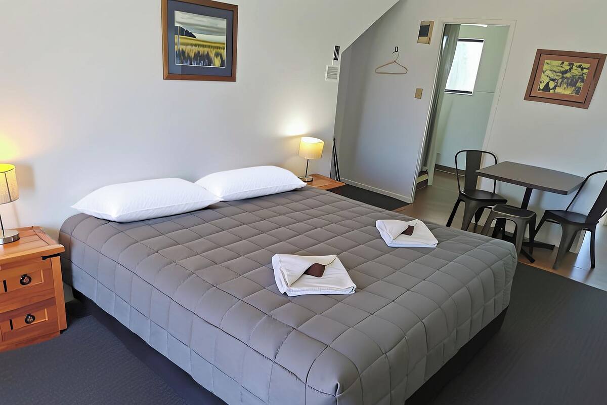 A neatly made bed with two pillows and two folded towels, a bedside table with a lamp, a small desk with a chair, and a framed picture on the wall.