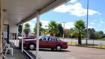 Un espacio al aire libre cubierto con un coche rojo aparcado cerca, sillas blancas y vistas a palmeras y un cielo despejado.