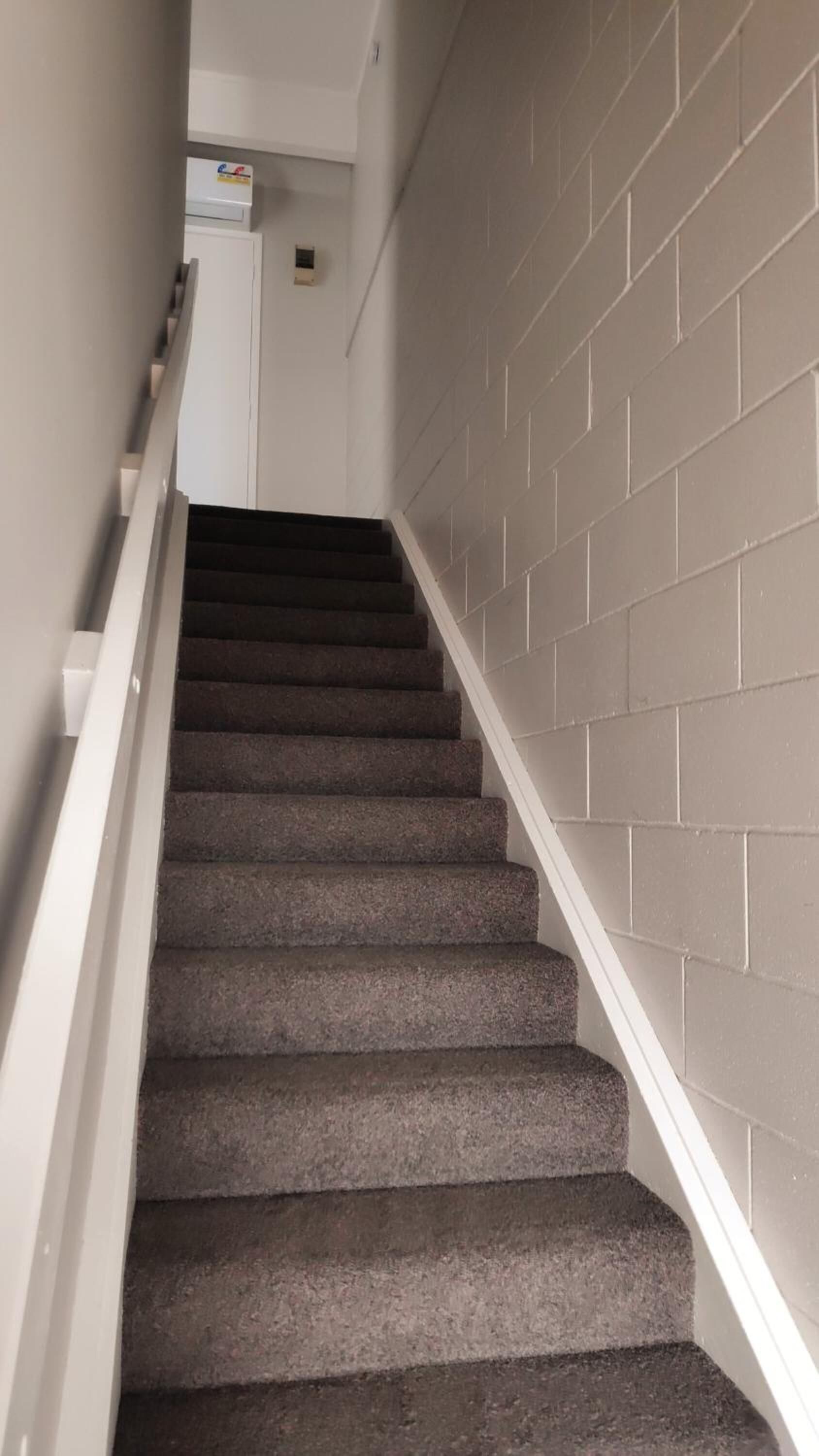 Un escalier avec des marches recouvertes d’une moquette sombre et des rampes blanches, menant à un palier avec un mur blanc et un petit objet blanc accroché au mur.