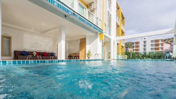 An indoor swimming pool with clear blue water, a sun terrace with a sofa and cushions, and a view of surrounding buildings. - Triple Three Patong (Patong)