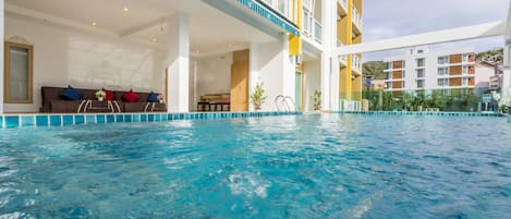 Una piscina interior con agua cristalina y azul, una terraza con sofás y cojines, y vistas de edificios circundantes.