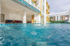 Une piscine intérieure à l’eau cristalline, une terrasse ensoleillée avec un canapé et des coussins, et une vue sur les bâtiments environnants.