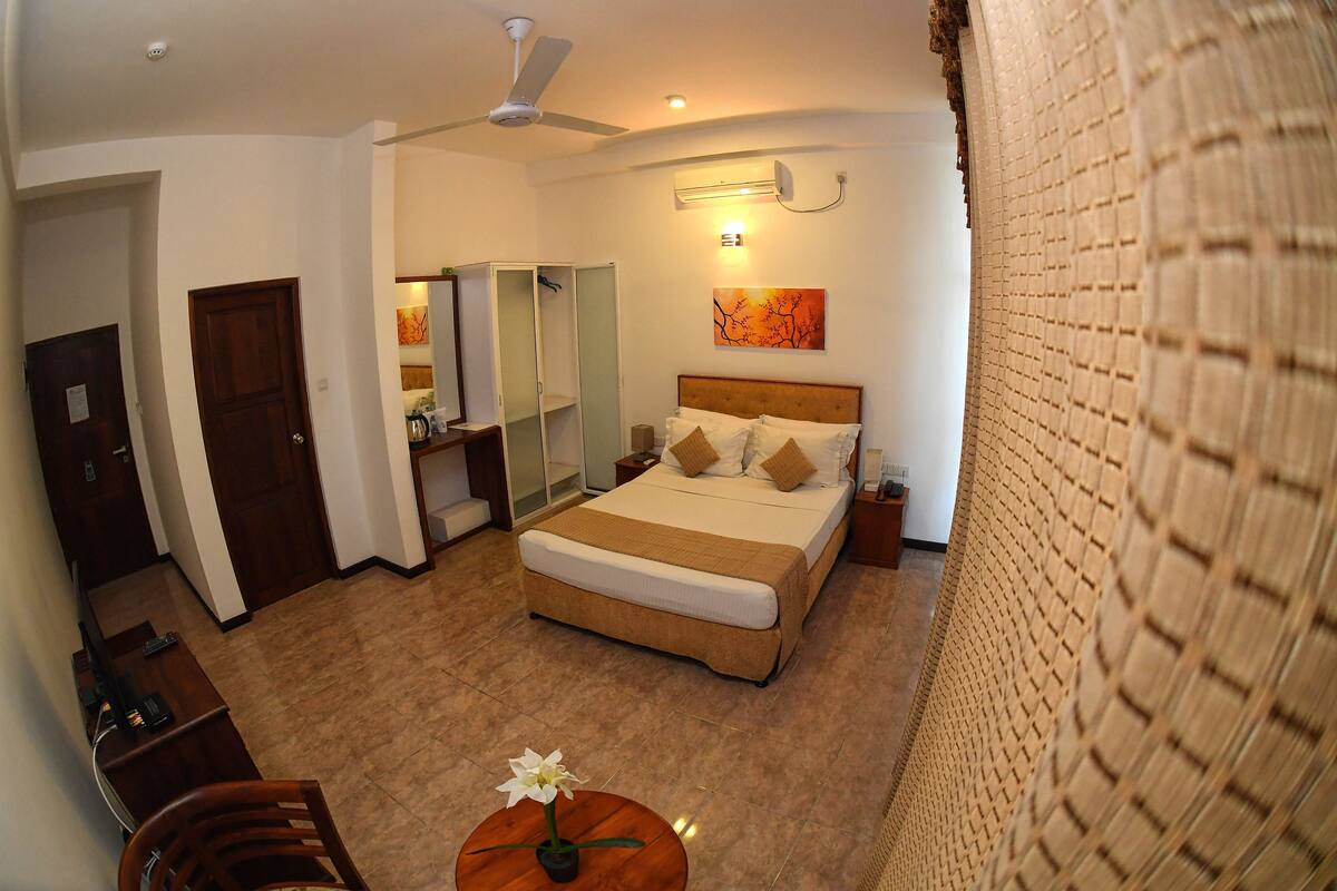 A hotel room with a bed, a desk, a chair, a round table with a flower, a ceiling fan, and a wall-mounted air conditioner.