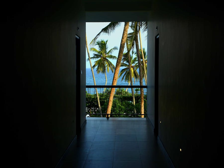 A hallway leading to a balcony with a view of palm trees and the ocean.