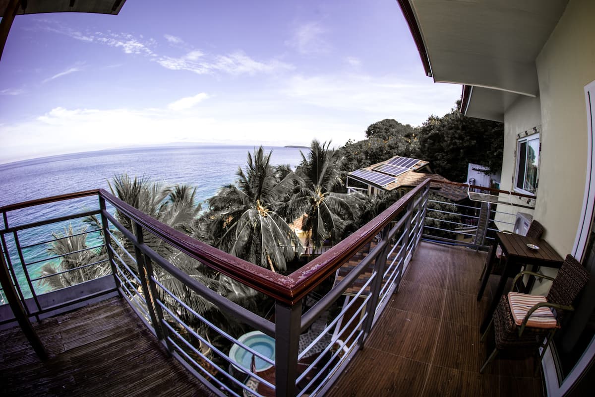 A balcony with a view of the ocean, palm trees, and a dining area with chairs and a table.