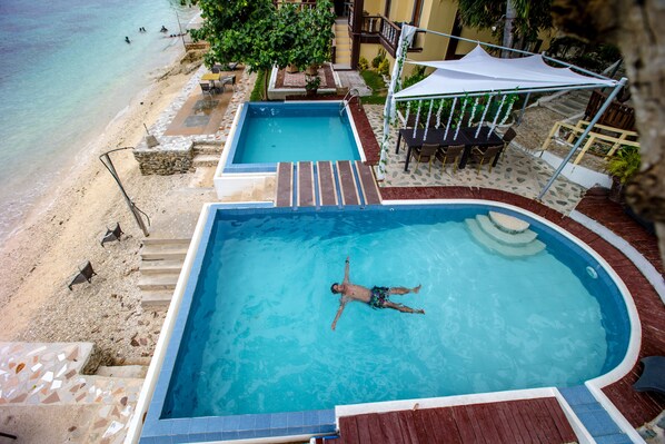 Una piscina con una persona flotando, rodeada por una terraza con una mesa y sillas, y vistas a la playa.