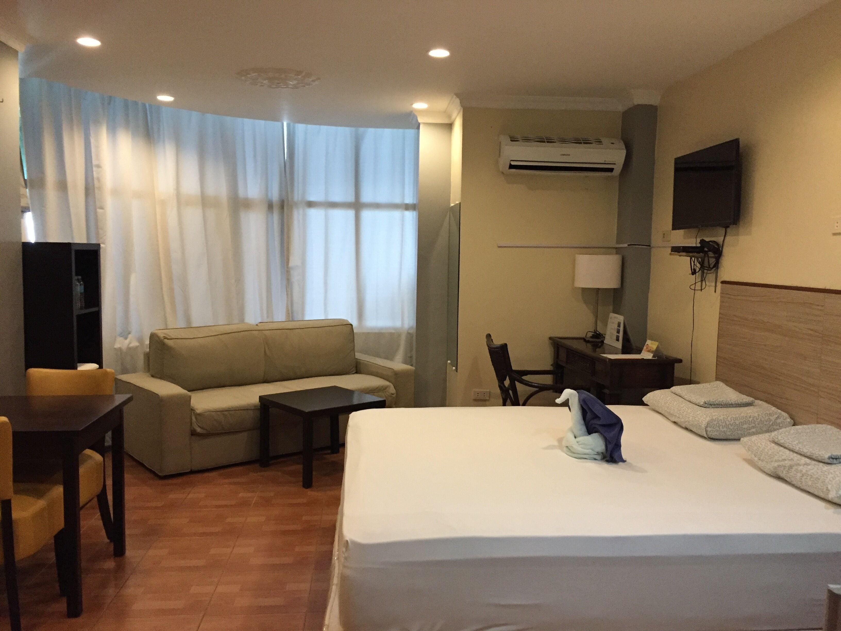 A hotel room with a bed, a sofa, a desk, a chair, an air conditioning unit, and a TV on the wall.