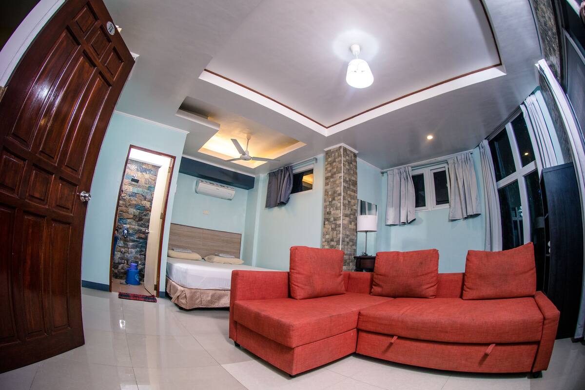 A room with a wooden door, a red sofa, a bed with a wooden headboard, a ceiling fan, and a window with curtains.