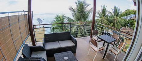 A balcony with wicker furniture, a table, and a view of the ocean and palm trees.