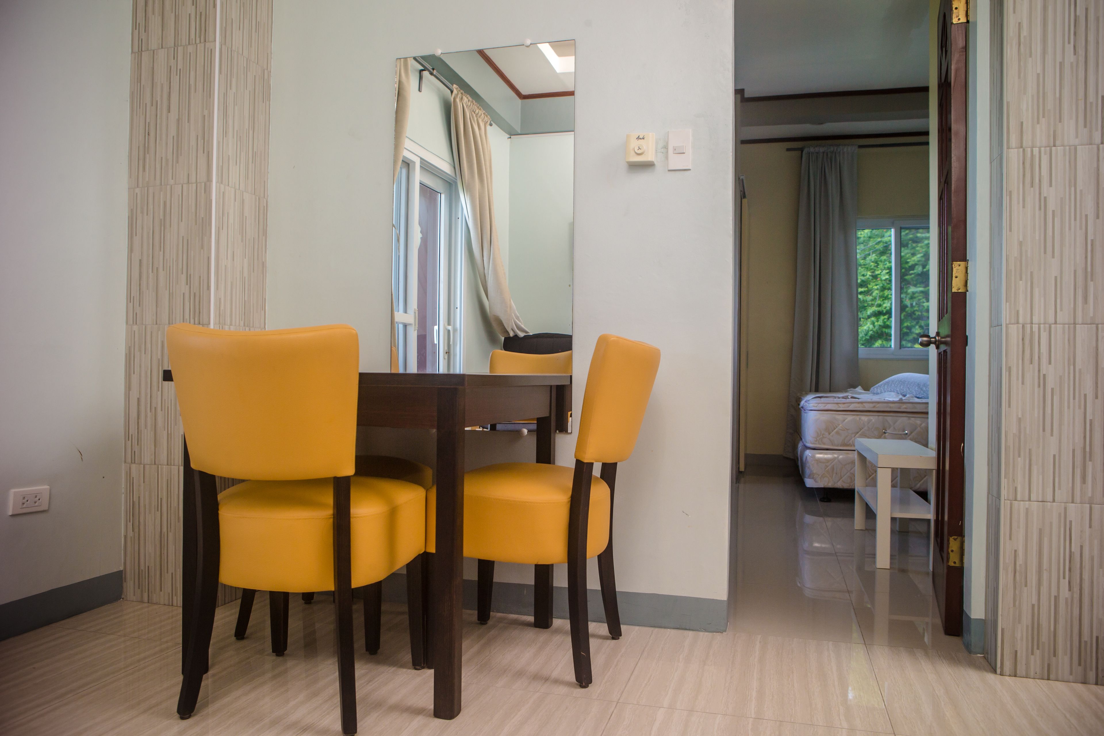 Een eetgedeelte met een tafel en vier gele stoelen, een slaapkamer met een bed en een raam, en een deur die leidt naar een andere kamer.