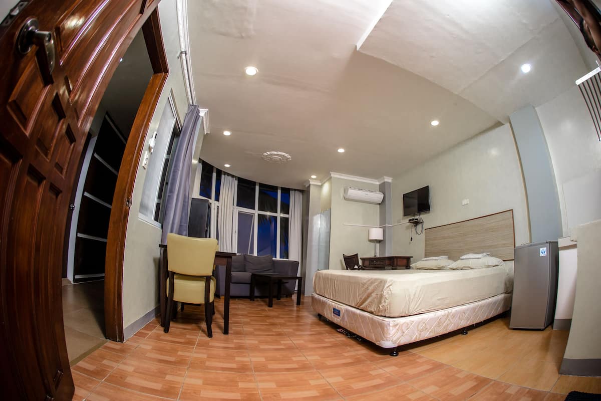 A hotel room with a bed, a desk, a chair, a TV, and an air conditioning unit.