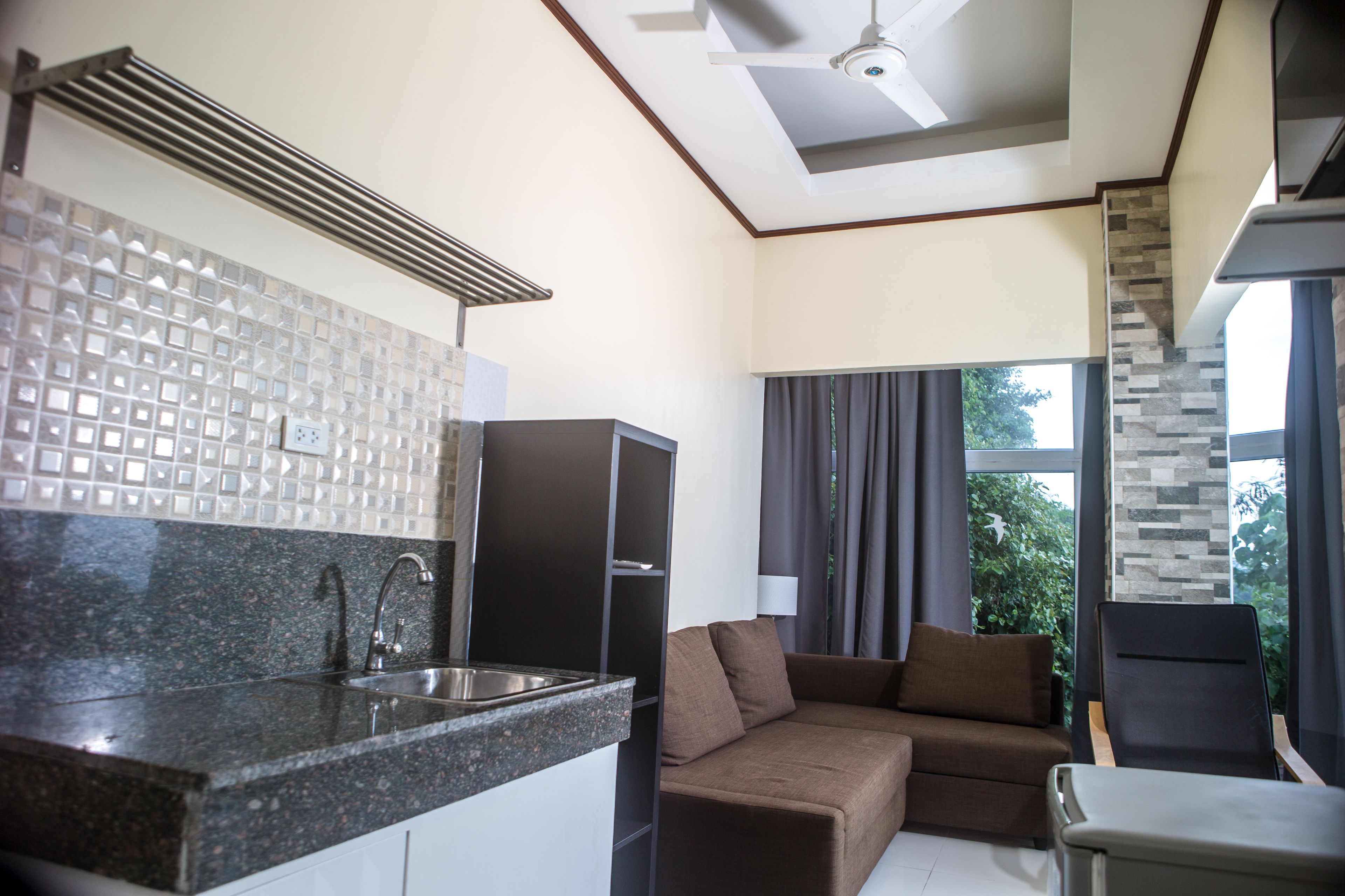 A modern kitchen with a granite countertop and stainless steel sink, a brown sofa, and a view of greenery through large windows.