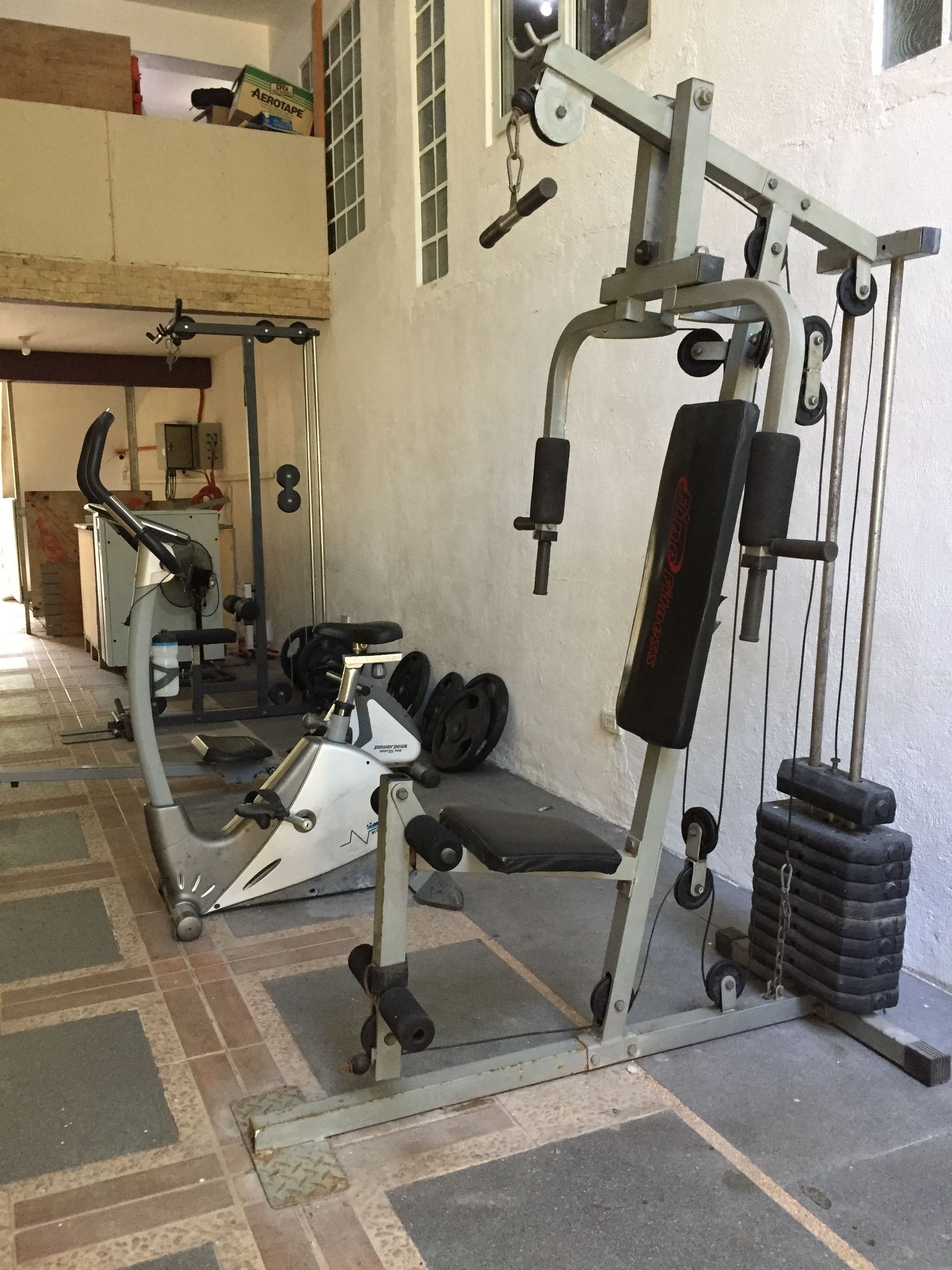 Un gimnasio con diverso equipo de ejercicio, incluyendo una prensa de banca, mancuernas y discos de pesas.