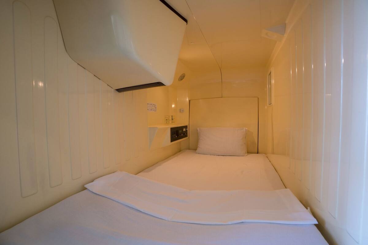 A small, single-bed room with a white bedspread, a headboard, and a wall-mounted control panel.