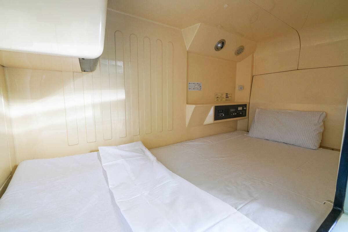 A small, single-bed room with a control panel, a window, and a radiator.