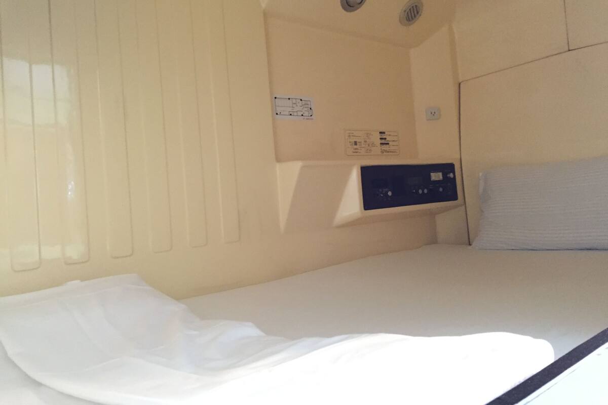 A compact, single-bed room with a control panel on the wall, a window, and a ceiling vent.