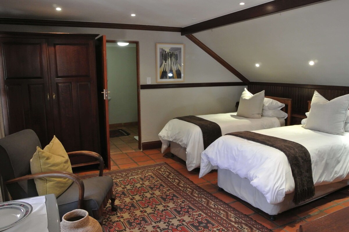 A bedroom with two single beds, a wooden door, a chair, a rug, and a painting on the wall.