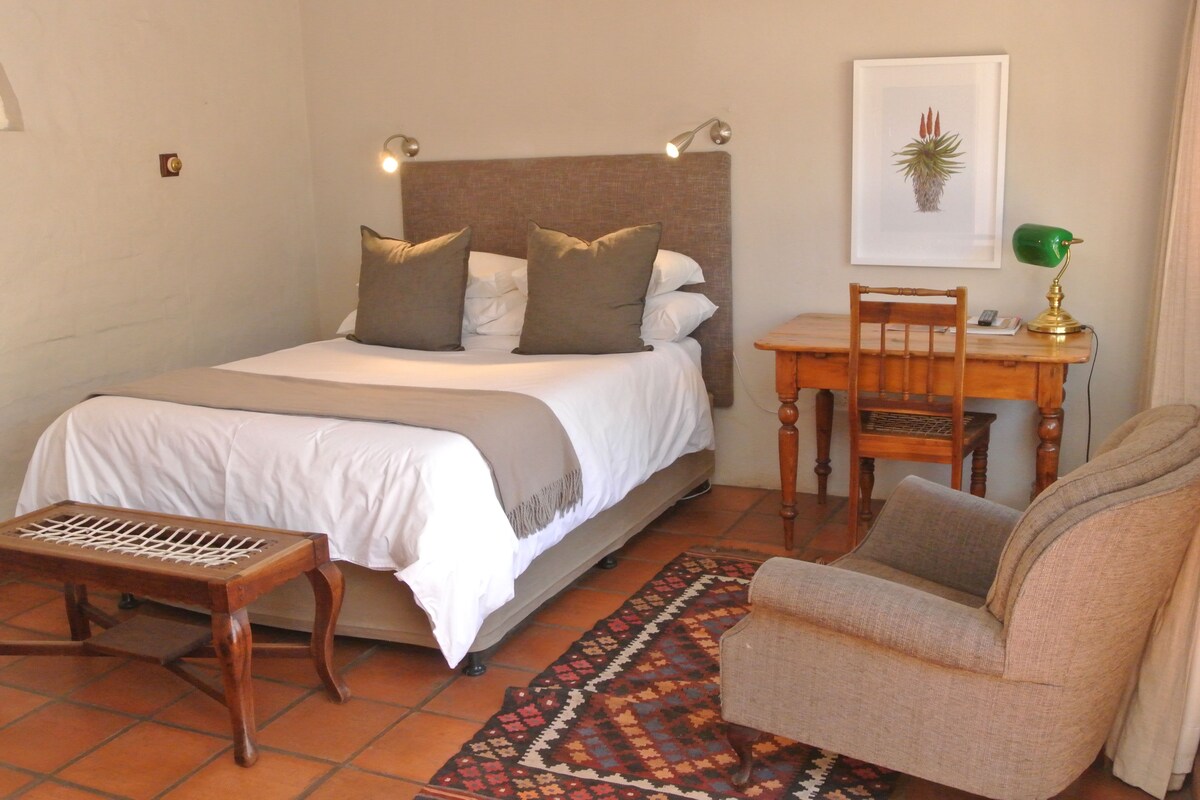 A bedroom with a bed, bedside table, chair, desk, lamp, and a framed picture on the wall.