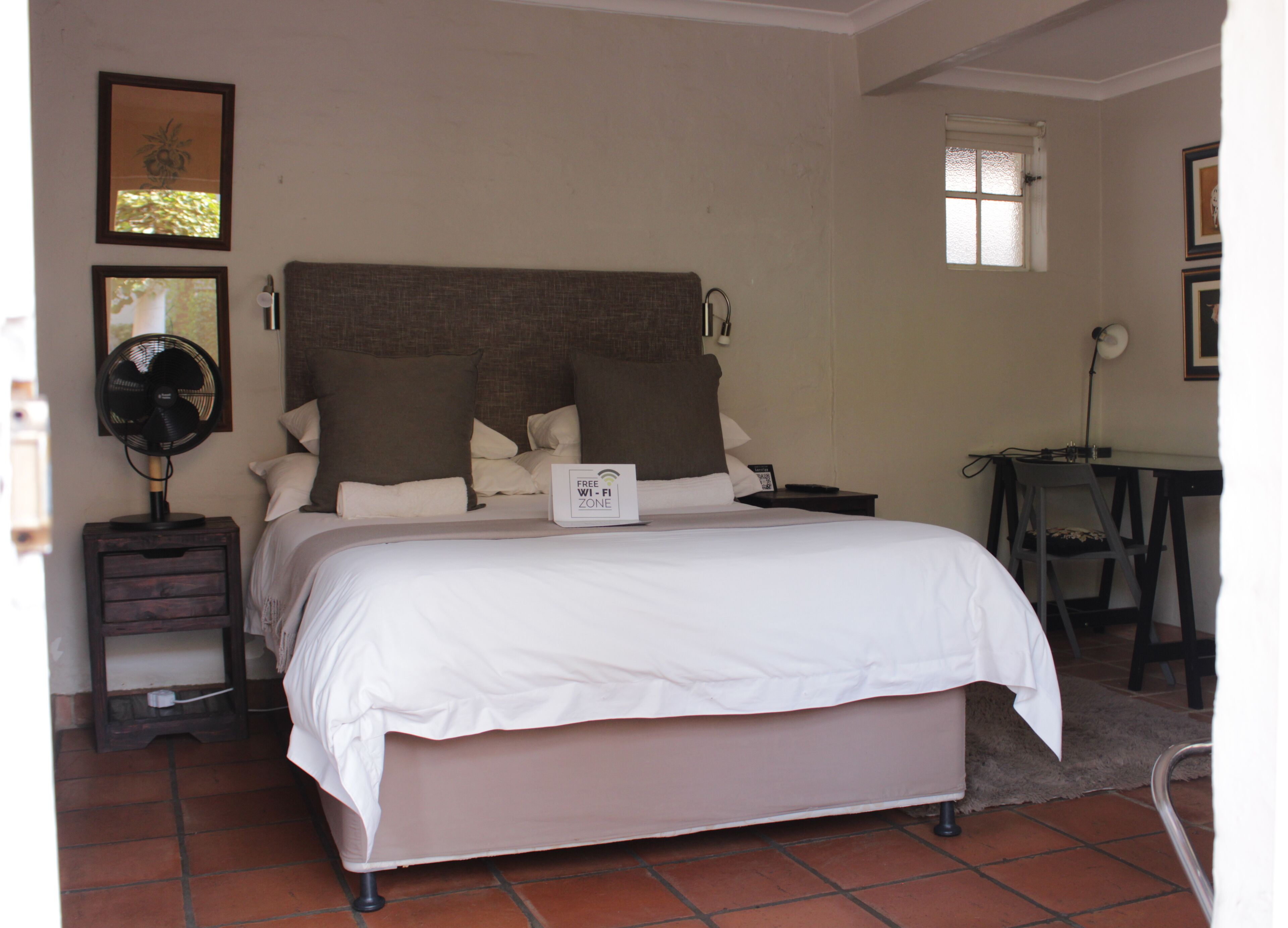A neatly made bed with a sign indicating a free Wi-Fi zone, a bedside table with a fan, and a small desk with a chair.