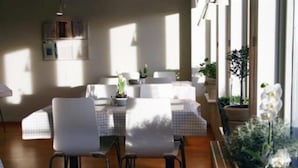 Un coin repas avec une table dressée pour quatre personnes, entourée de chaises blanches, agrémenté de plantes en pot et offrant une vue sur l’extérieur.