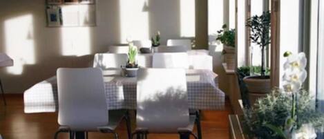 A dining area with a table set for four, surrounded by white chairs, with potted plants and a view of the outdoors.