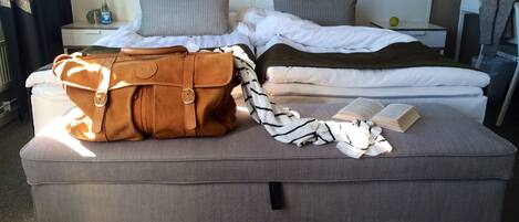 A neatly made bed with pillows, a brown leather bag, a striped blanket, and an open book on a grey ottoman in a hotel room.