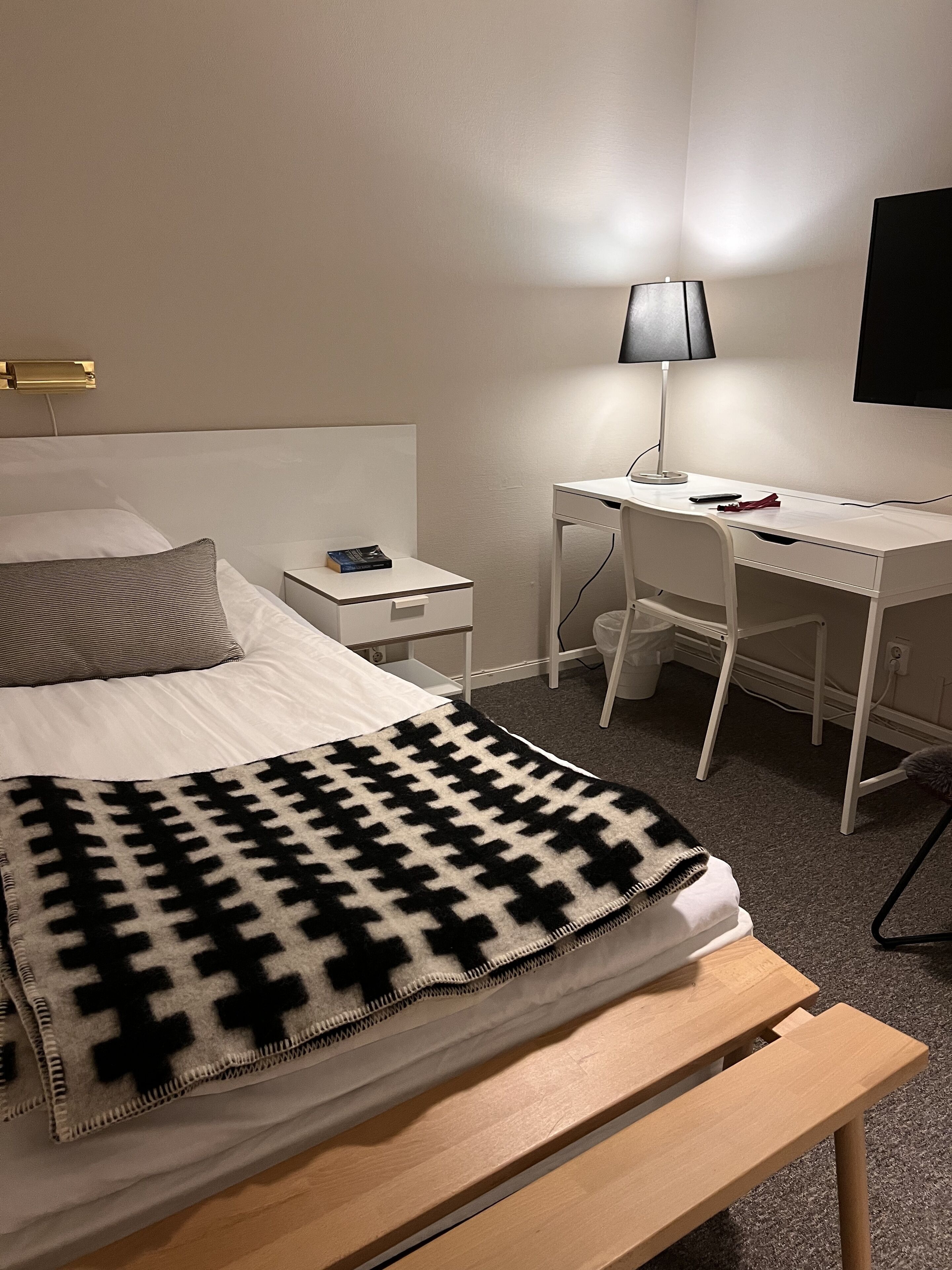 A hotel room with a bed, a desk, a chair, a lamp, and a TV.