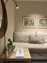 Una habitación con una mesa de madera, un florero de vidrio con una planta, un cuadro enmarco y un sofá con cojines.