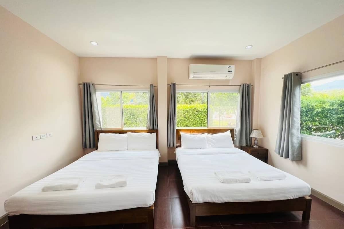A hotel room with two beds, a window with curtains, an air conditioning unit, and a view of greenery outside.