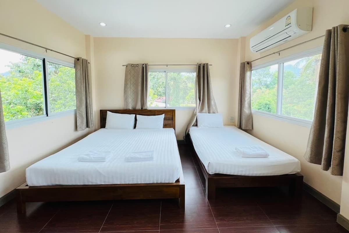 A room with two single beds, a wooden headboard, beige walls, and large windows with curtains.