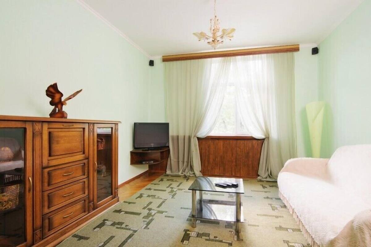 A room with a wooden cabinet, a television, a glass coffee table, a sofa, and a window with sheer curtains.
