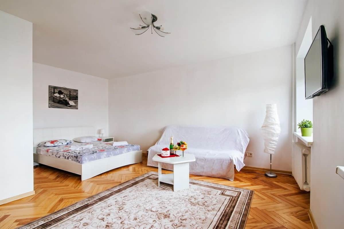 A modern, well-lit bedroom with a bed, bedside table, and a small table with a bottle and flowers.