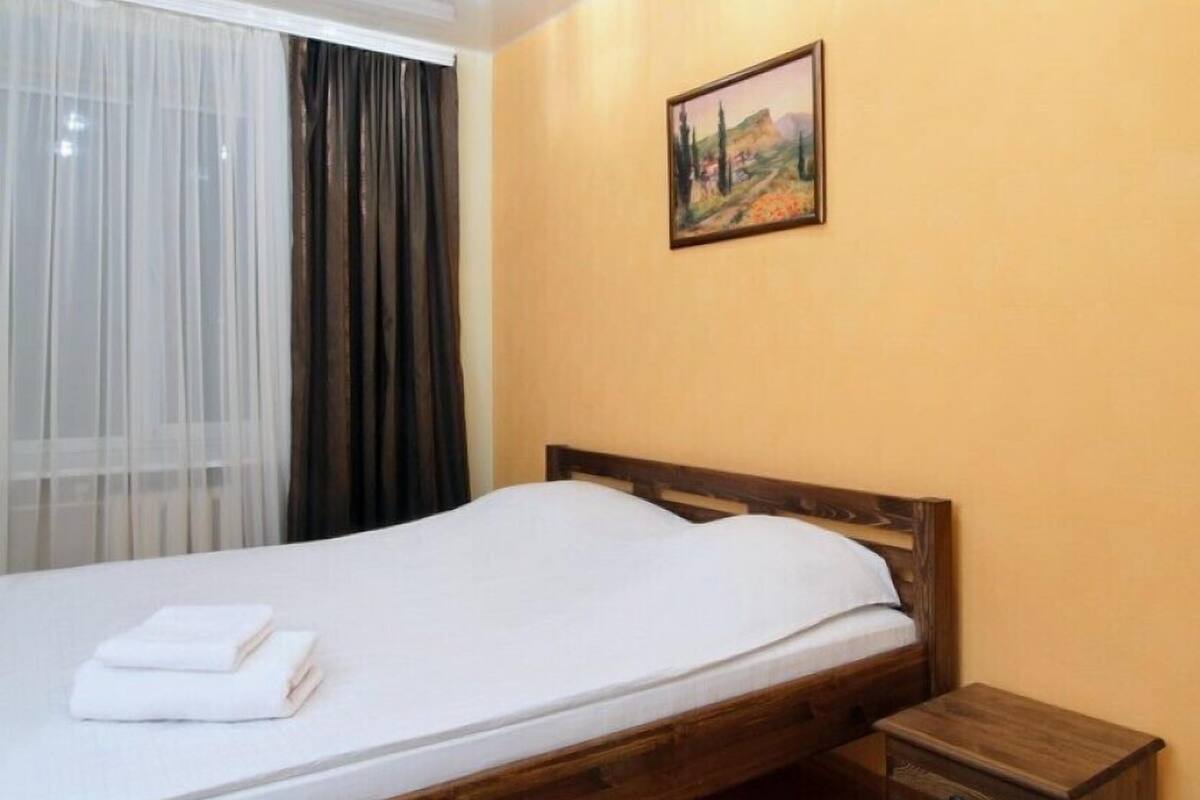 A bedroom with a wooden bed, a small bedside table, a nightstand, a framed picture on the wall, and a window with sheer curtains.