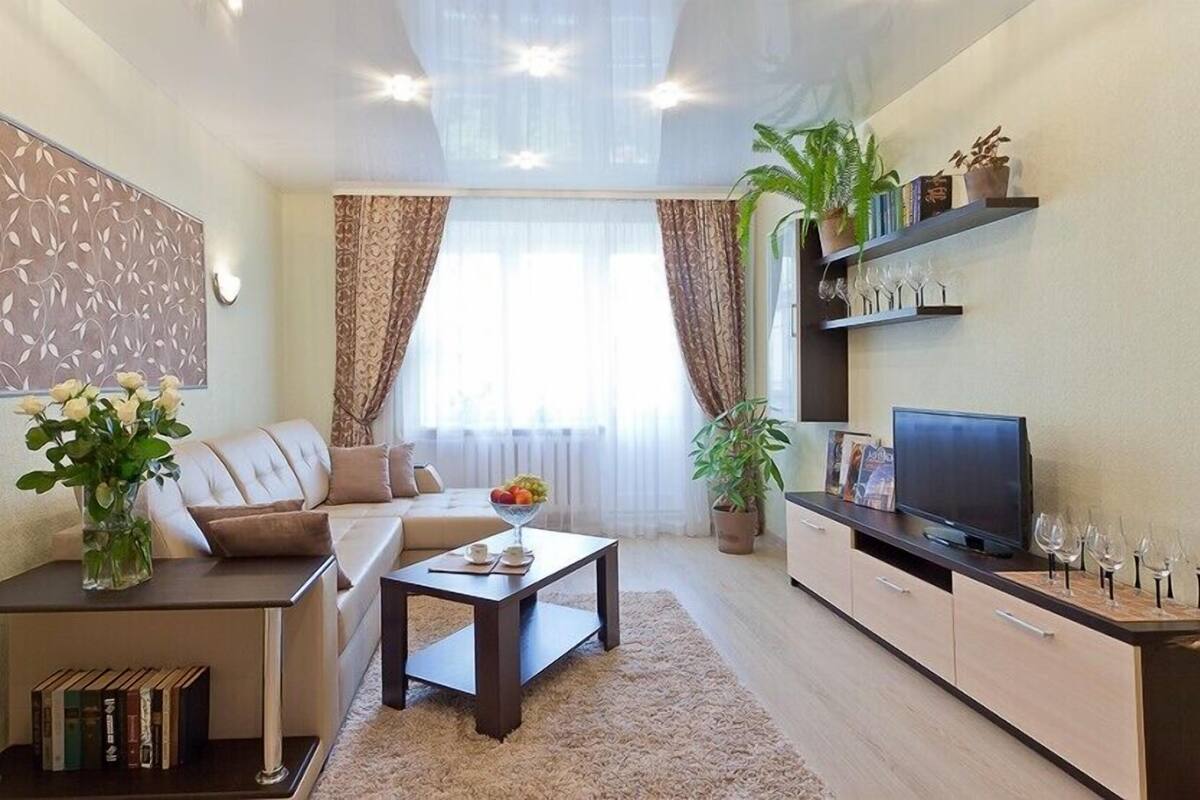 A modern living room with a sofa, coffee table, and TV stand. There are books, a vase with flowers, and a plant on the shelves.
