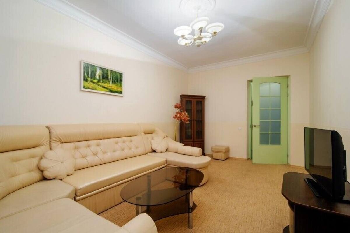 A living room with a beige sectional sofa, a glass coffee table, a television on a stand, a green door, and a painting on the wall.