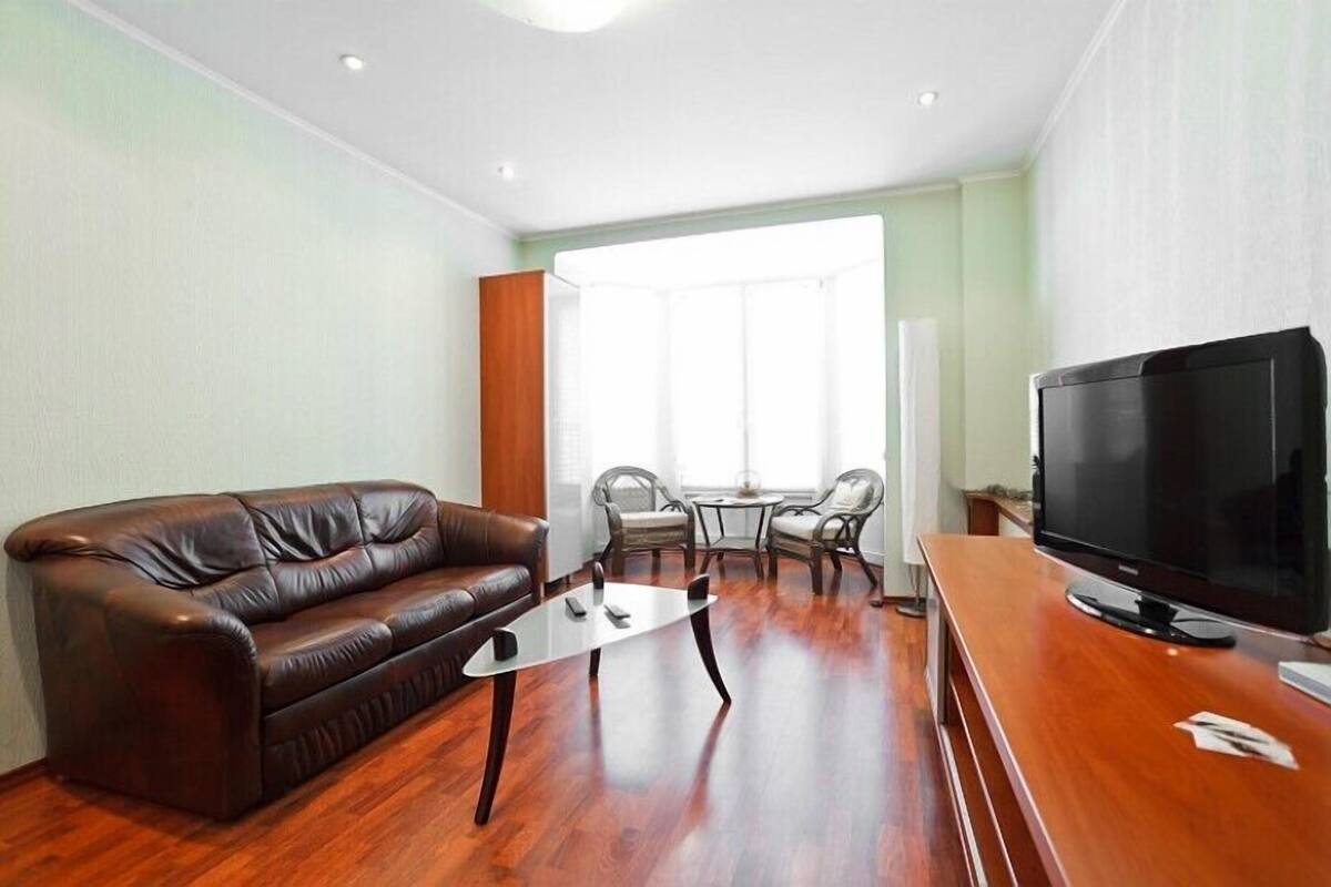 A living room with a brown leather sofa, a glass coffee table, a TV on a wooden stand, and a dining area with chairs and a table.