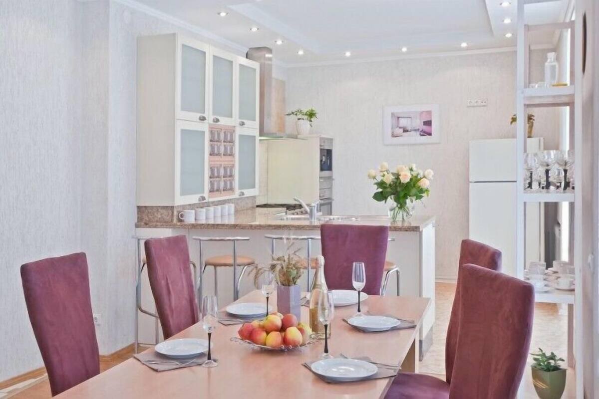 A dining area with a long table, red chairs, and a fruit bowl. There are white cabinets and a white refrigerator in the background.