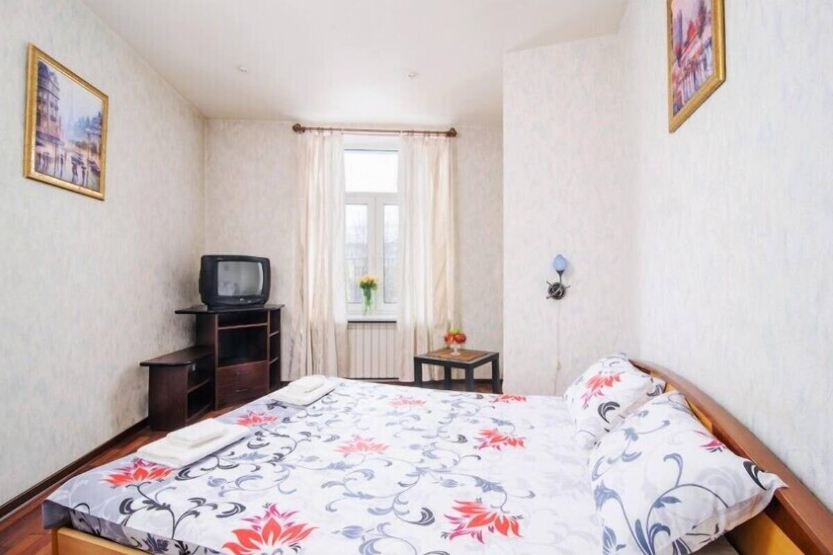 A bedroom with a bed, a television on a stand, a window with curtains, and a small table with a vase.