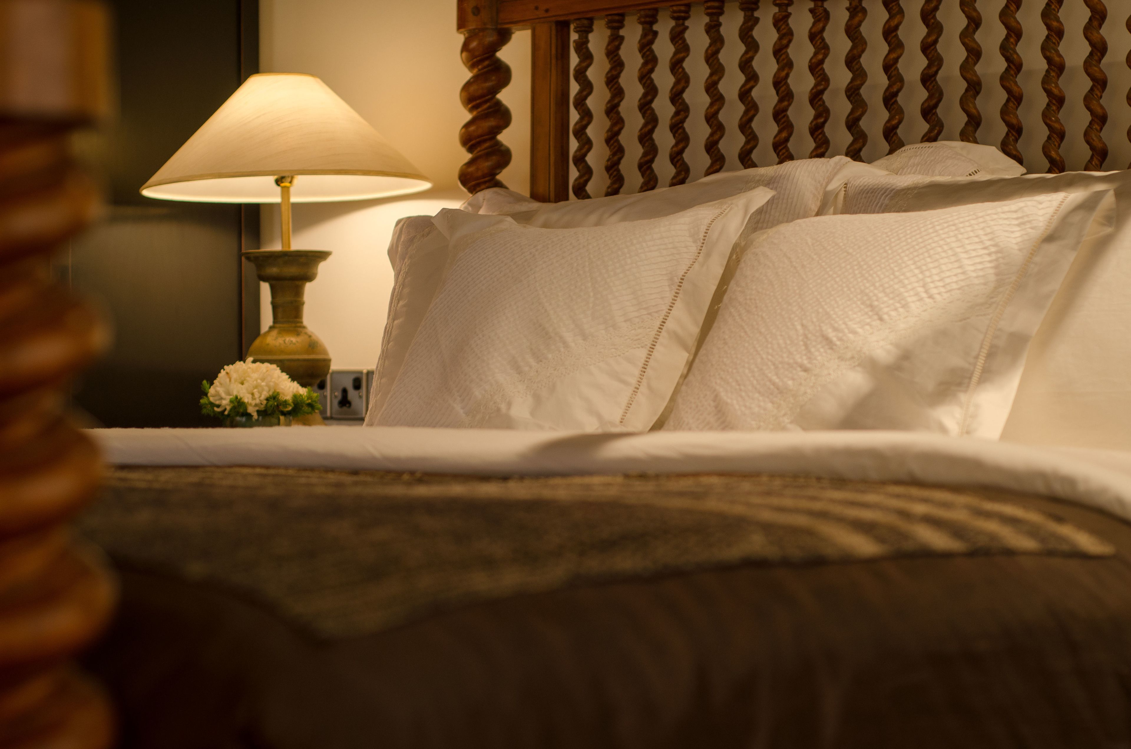 A neatly made bed with white linens, a wooden headboard, and a bedside lamp with a floral arrangement.