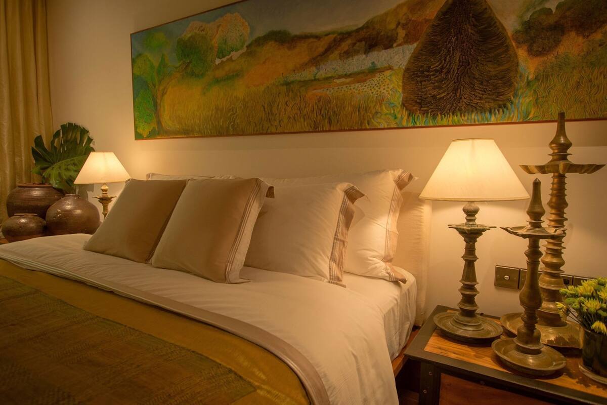 Habitación de hotel con cama, dos lámparas de noche, un cuadro en la pared y una mesita de noche con objetos decorativos.