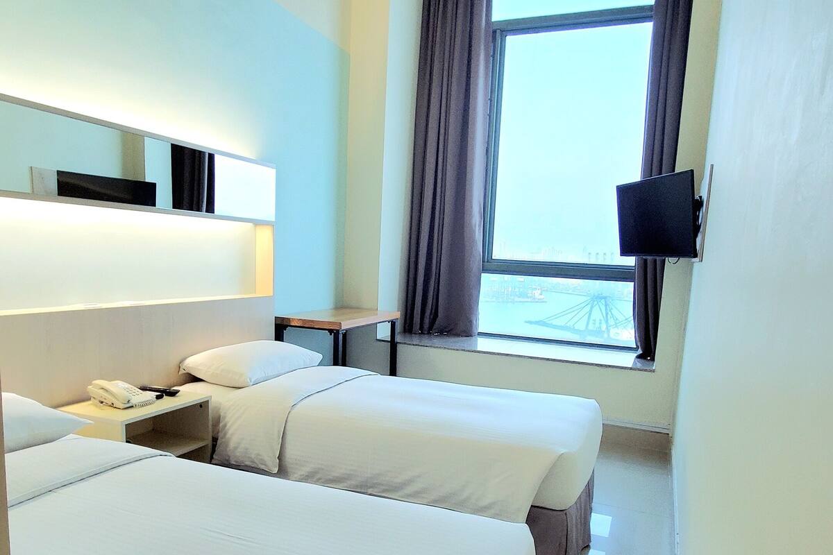 A hotel room with two beds, a bedside table with a phone, a flat-screen TV mounted on the wall, and a window with curtains.