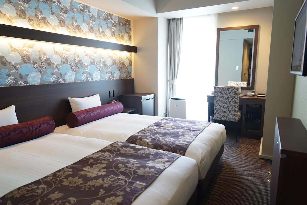 A hotel room with two beds, a desk, a mirror, and a bathroom.