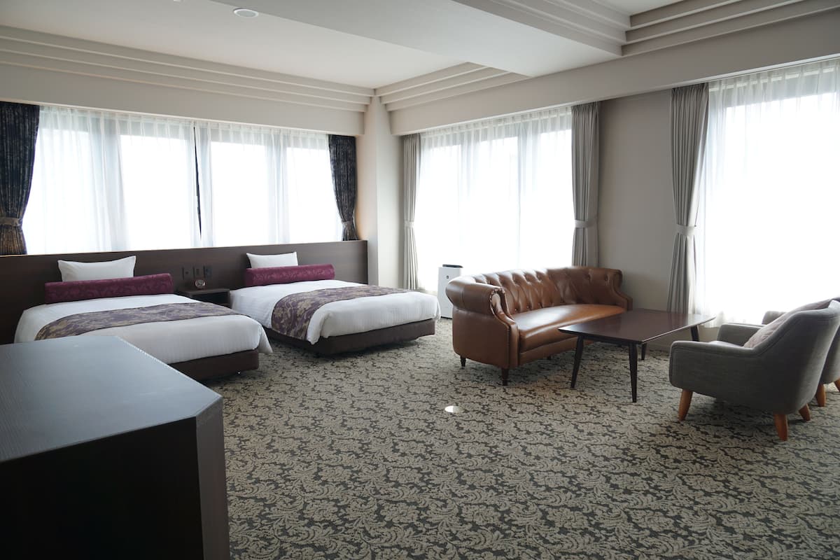 A hotel room with two beds, a sofa, a table, and large windows with curtains.
