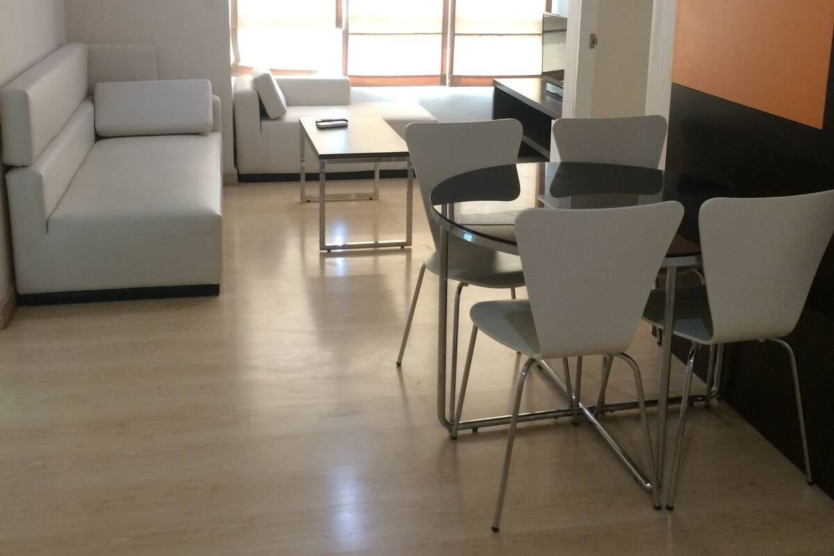 A modern living room with a sofa, a dining table with chairs, and a large window with blinds.