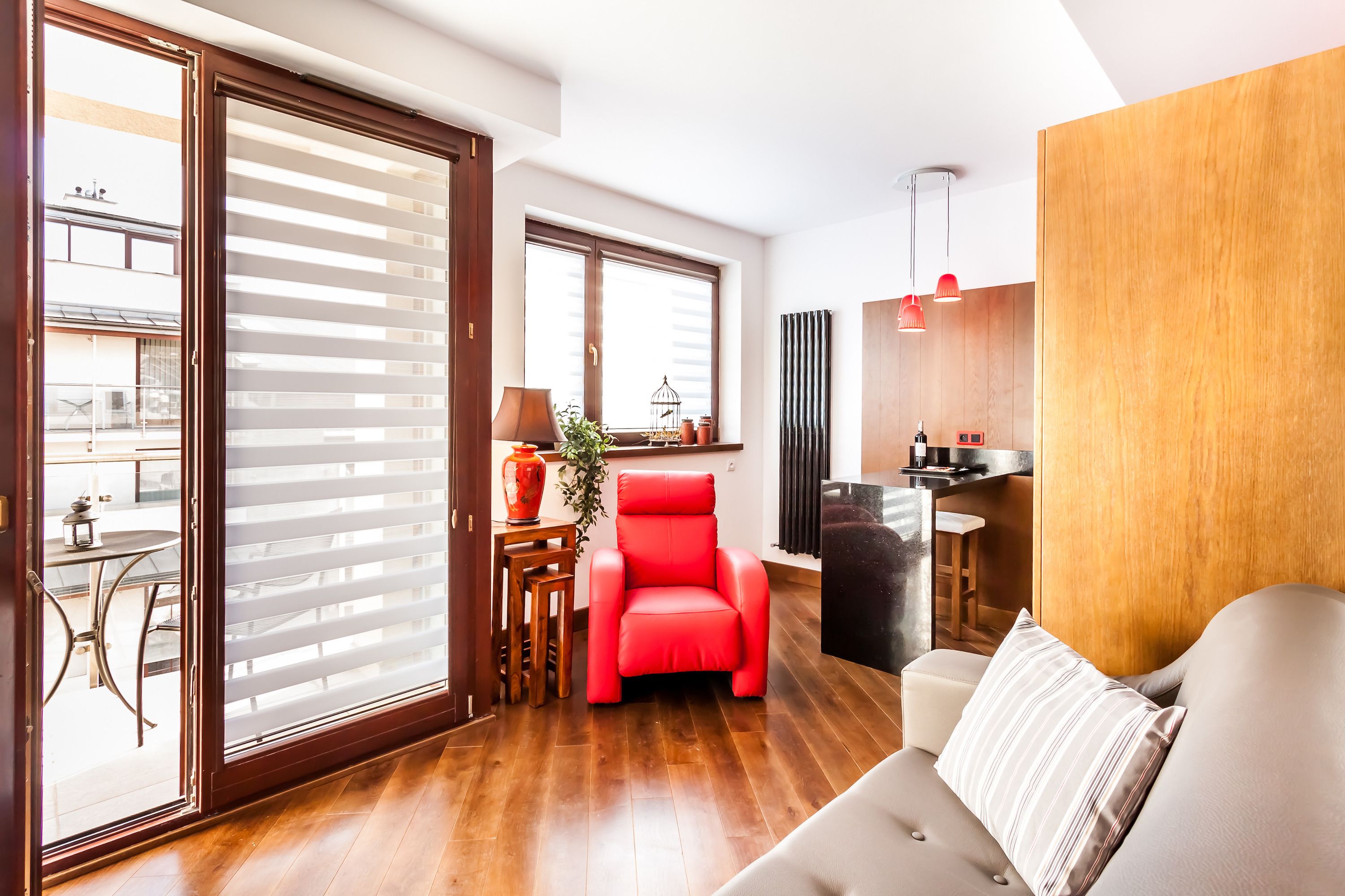 A modern living room with a red chair, a beige sofa, and a wooden floor. There is a sliding glass door leading to a balcony, and a kitchen area with a sink and countertop.