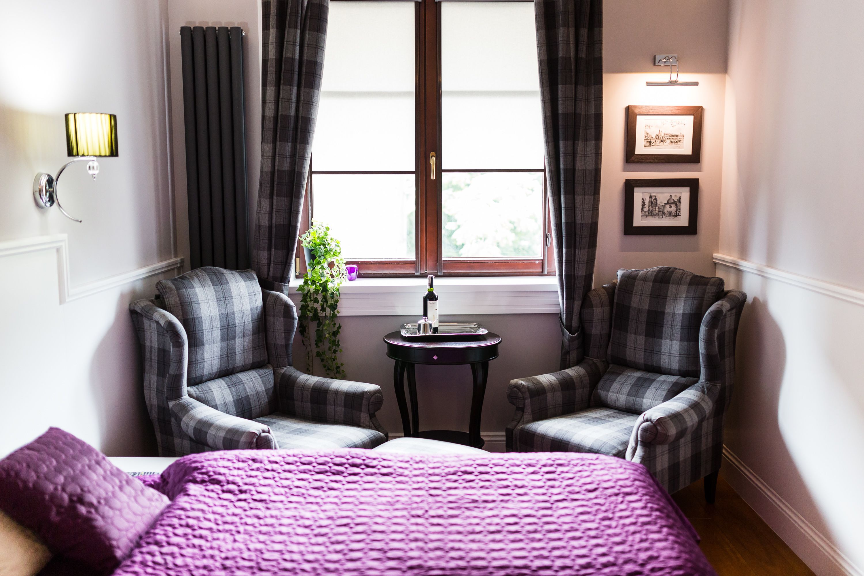 Een kamer met een bed, twee fauteuils, een tafeltje, een raam met gordijnen, en wandlampen.